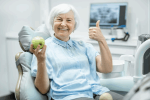 Woman in patient’s chair smiling and giving a thumbs up while holding an apple